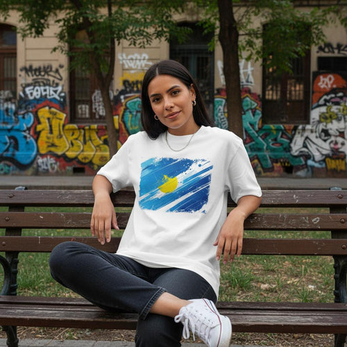 Argentina Albiceleste Light Mode. Woman sitting on a bench wearing a white t-shirt with a blue graphic design, in an urban setting with graffiti.