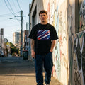 Australia Socceroos Dark. Person wearing a black t-shirt with a colorful graphic design, standing on a street with graffiti-covered walls.