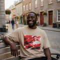 Canada Les Rouges Sand. Man sitting on a bench wearing a beige t-shirt with a red graphic design, smiling in an urban setting.