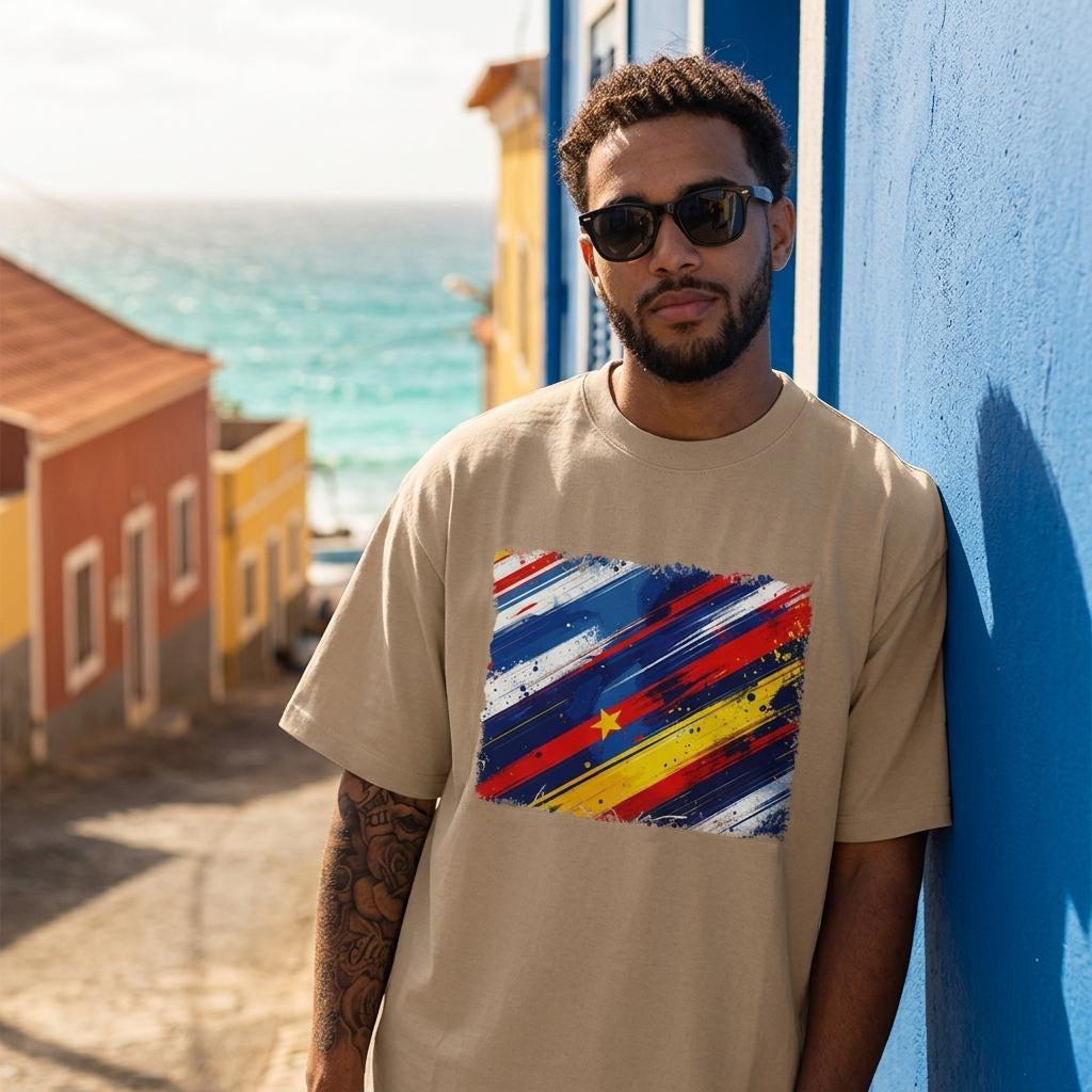 Cape Verde Tubaroes Azules Sand. Man wearing a beige t-shirt with a colorful abstract design, standing against a blue wall with ocean view.