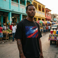 Haiti Les Grenadiers Dark. Man wearing a black t-shirt with a colorful graphic design in an outdoor market setting