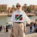 Iraq Lions of Mesopotamia Light. Woman wearing a white t-shirt with a colorful design and beige hijab in an outdoor setting with water and boats.