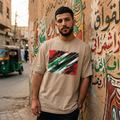 Iraq Lions of Mesopotamia Sand. Man wearing a beige t-shirt with a colorful design, standing against a wall with Arabic text and graffiti.