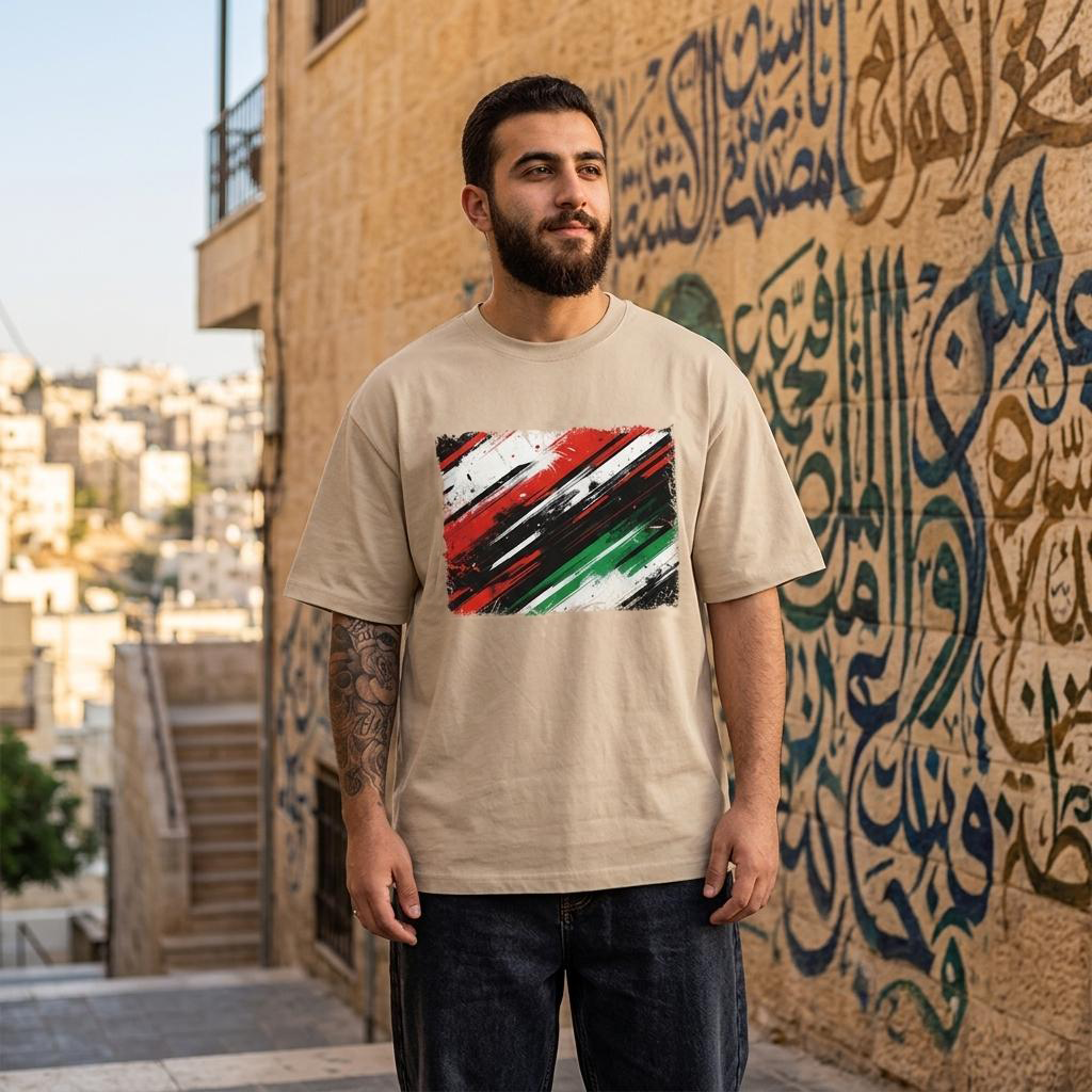 Jordan Nashama Sand. Man wearing a beige t-shirt with a colorful design in front of a textured wall with Arabic calligraphy.