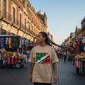 Mexico El Tri Sand Mode. Woman wearing a beige t-shirt with a colorful graphic design in an outdoor market setting.