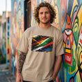 South Africa Bafana Bafana Sand Man wearing a beige t-shirt with a colorful abstract design, standing in front of a vibrant mural.